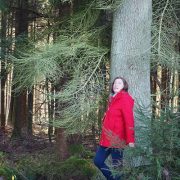 Rotjäckchen im Wald: Frau mit rotem Jäckchen steht angelehnt an einem Baum und schaut in die Ferne.