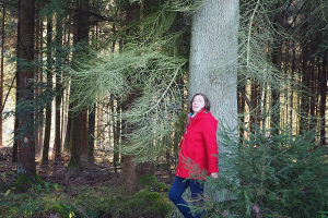 Rotjäckchen im Wald: Frau mit rotem Jäckchen steht angelehnt an einem Baum und schaut in die Ferne.