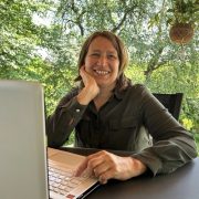 Anita sitzt mit ihrem Laptop im Garten und arbeitet konzentriert, umgeben von natürlichem Grün.