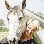 Claudia Friederich lacht glücklich neben ihrem weißen Pferd, während sie sich an dessen Hals lehnt.