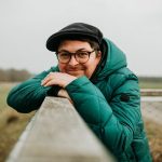 Porträt eines Mannes mit Brille, grüner Jacke und Mütze, der lächelnd an einem Geländer im Freien lehnt – Kundenstimme zum Emotions‑Coaching.