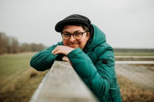 thomas-kern.jpg Porträt eines Mannes mit Brille, grüner Jacke und Mütze, der lächelnd an einem Geländer im Freien lehnt – Kundenstimme zum Emotions‑Coaching.