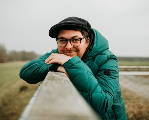 Porträt eines Mannes mit Brille, grüner Jacke und Mütze, der lächelnd an einem Geländer im Freien lehnt – Kundenstimme zum Emotions‑Coaching.