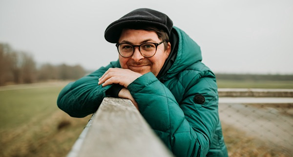 Porträt eines Mannes mit Brille, grüner Jacke und Mütze, der lächelnd an einem Geländer im Freien lehnt – Kundenstimme zum Emotions‑Coaching.