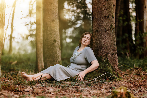 Anita Griebl liegt im Wald an einem Baumstamm und hat die Augen geschlossen – Symbol für innere Blockaden, Erschöpfung und den Weg zurück ins Fühlen.