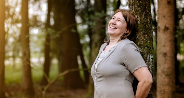 Anita Griebl im Wald, an einen Baum gelehnt, mit weitem Blick – Moment der Einkehr, Energiearbeit und intuitiven Selbstverbindung