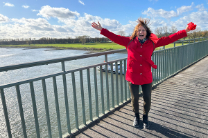 Anita Griebl springt auf einer Brücke, trägt eine rote Jacke, der Wind weht ihr die Haare ins Gesicht – Moment voller Leichtigkeit.