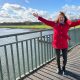 Anita Griebl springt auf einer Brücke, trägt eine rote Jacke, der Wind weht ihr die Haare ins Gesicht – Moment voller Leichtigkeit.
