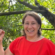 „Anita Griebl lächelt in der Natur in die Kamera und hebt beide Hände in einer offenen, einladenden Geste.