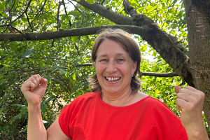 Fühle Energie. Lebe frei „Anita Griebl lächelt in der Natur in die Kamera und hebt beide Hände in einer offenen, einladenden Geste.