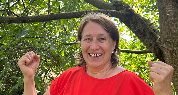 „Anita Griebl lächelt in der Natur in die Kamera und hebt beide Hände in einer offenen, einladenden Geste.