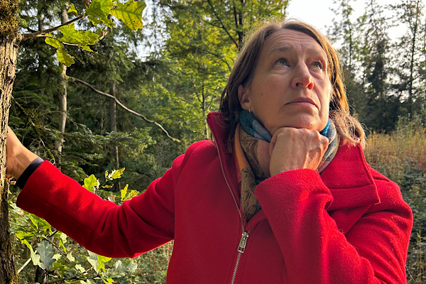 Beitragsbild zum Abschnitt Spiritualität geht online: Anita Griebl in roter Jacke im Wald schaut zum Himmel.