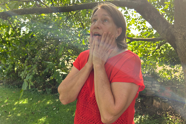 Anita Griebl steht unter einem Baum in der Natur, trägt ein rotes Shirt, schaut nach oben und hält ihr Gesicht sanft in den Händen, während ein Lichtstrahl zwischen den Ästen hindurchfällt.