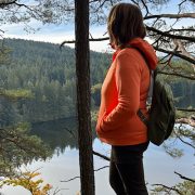 „Anita Griebl mit Rucksack steht inmitten der Natur und blickt auf einen ruhigen See, umgeben von Bäumen – Symbol für Innenschau, emotionale Klarheit und Tools für Veränderungsfreudige
