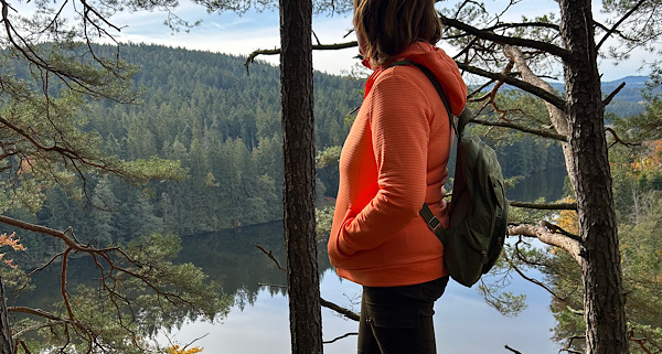 „Anita Griebl mit Rucksack steht inmitten der Natur und blickt auf einen ruhigen See, umgeben von Bäumen – Symbol für Innenschau, emotionale Klarheit und Tools für Veränderungsfreudige