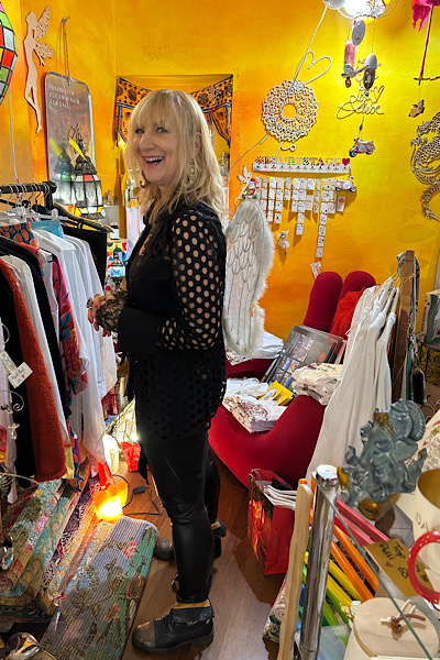 Gabi Oberberger steht seitlich im Feng‑Shui‑Laden, trägt schwarze Kleidung und weiße Engelsflügel; im Hintergrund sind bunte Waren und Dekorationen zu sehen.