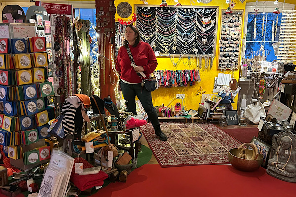 Heidi steht vor dem Konplott‑Schmuckregal; im Hintergrund ist eine große Auswahl an bunten Waren aus dem Feng‑Shui‑Laden zu sehen.