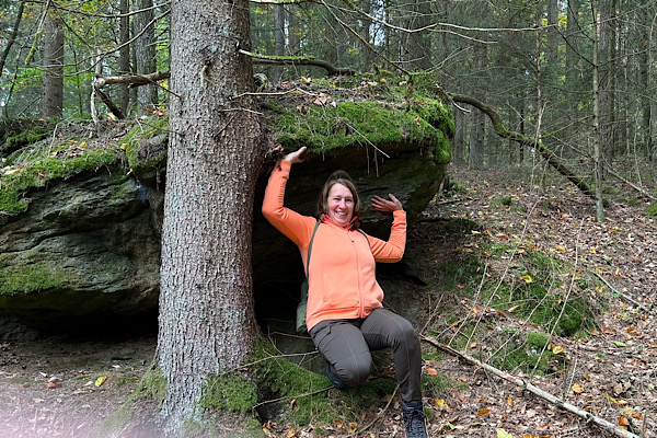 Anita Griebl in oranger Jacke hockt im Wald unter einem großen Stein und tut so, als würde sie ihn hochheben – Symbol für mentale Last und Gedanken, die schwer wirken.