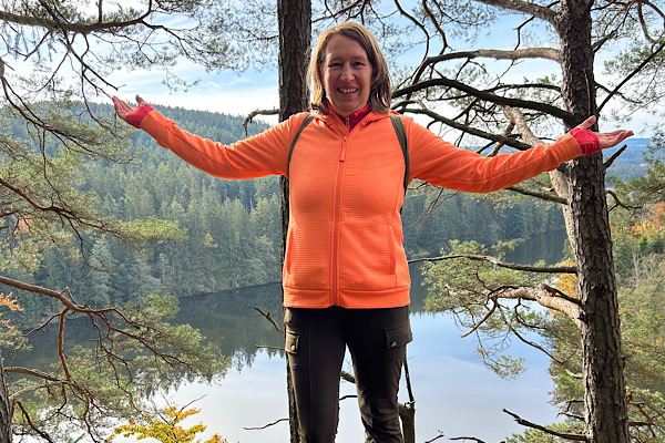 Anita Griebl in oranger Jacke steht im Wald mit ausgebreiteten Armen vor einem See – Symbol für Heilenergetik und Naturverbundenheit.