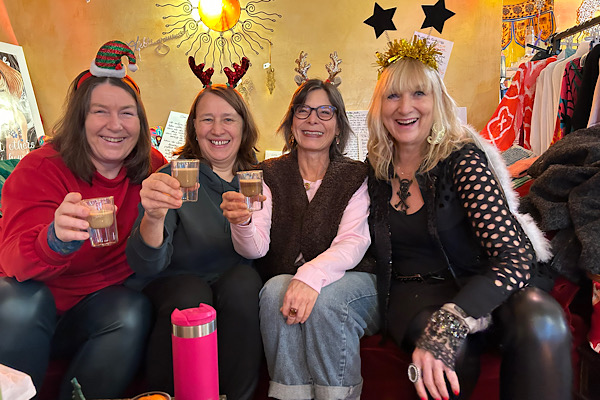Vier Frauen sitzen auf einer Couch, tragen weihnachtlichen Kopfschmuck und halten Tassen mit Rohkakao; im Hintergrund ist der Laden zu sehen.
