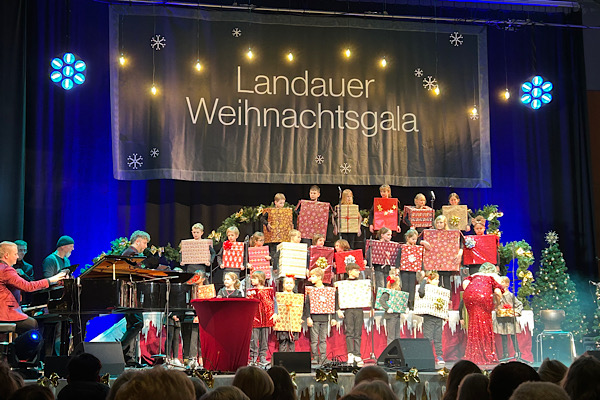 Bühne einer Weihnachtsgala mit Musikern und vielen Kindern, die als Geschenk‑Schachteln verkleidet sind.