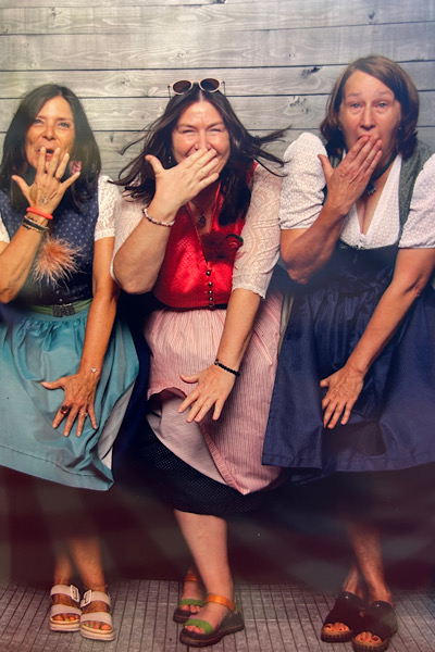 Drei Frauen in Dirndl beugen sich nach vorne und halten schamhaft eine Hand vor den Mund.