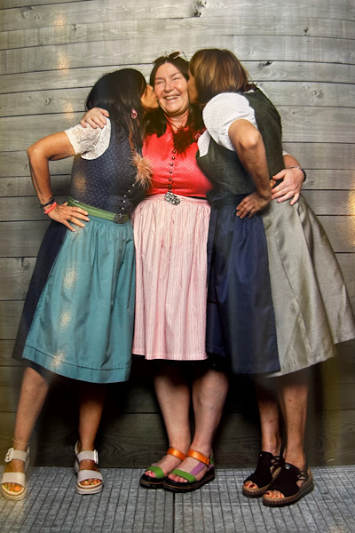 Zwei Frauen küssen eine lachende dritte Frau auf die Wange; alle haben die Arme in die Hüfte gestützt.