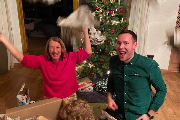 Anita Griebl und Alexander packen Weihnachtsgeschenke aus und werfen lachend Geschenkpapier in die Luft; im Hintergrund steht der Christbaum.