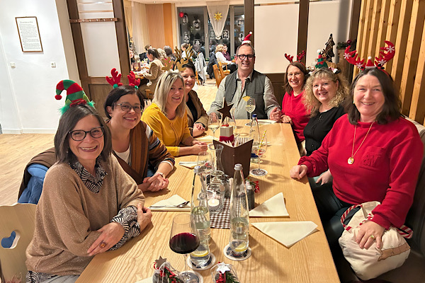 Sieben Frauen mit weihnachtlichen Haarreifen sitzen an einem Tisch; ein Kellner mit Nikolausmütze sitzt lächelnd in ihrer Mitte.