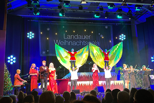 Bühne mit vielen Musikern und vier Personen, die eine akrobatische Übung zeigen: Zwei Männer halten jeweils ein Mädchen mit ausgebreiteten ‚Engelsflügeln‘ hoch in die Luft.