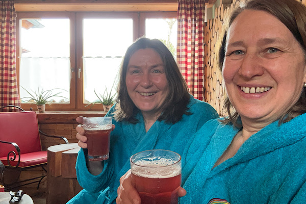 Anita Griebl und ihre Freundin Heidi im Wellnessbereich in blauen Bademänteln, beide halten ihr Energiedrink ‚Luminate‘ in den Händen.