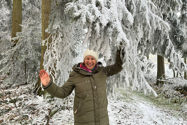 Anita Griebl zieht im winterlichen Wald an einem Fichtenzweig, sodass Schnee über ihr Gesicht rieselt.