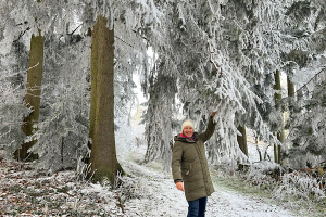 Mein Motto für 2026 Anita Griebl steht im winterlichen Wald und zieht an einem Fichtenzweig, sodass Schnee auf sie herabfällt.