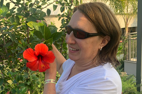 Anita mit Sonnenbrille hält eine rote Hibiskusblüte vor ihr Gesicht; ein naher, lebendiger Moment voller Farbe und Lebensfreude.