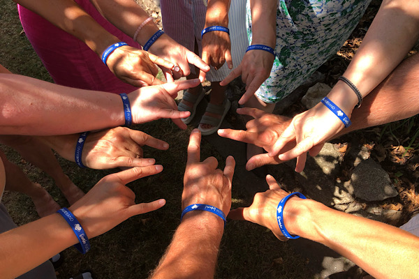 Mehrere Menschen führen ihre Hände im Kreis zusammen, verbunden durch blaue Bänder – ein Symbol für Gemeinschaft und Zusammenhalt.