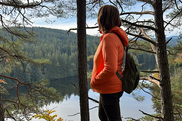 Anita Griebl steht mit Rucksack im Wald, dreht sich seitlich und blickt in die Ferne, während sie über alte Emotionen und inneres Loslassen reflektiert.