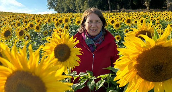 Anita Griebl steht in einem Feld voller Sonnenblumen und strahlt Ruhe, Präsenz und Lebensfreude aus.