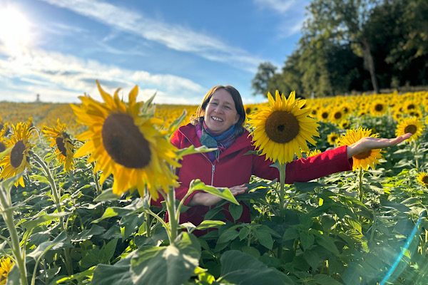 Anita steht zwischen hohen Sonnenblumen und streckt beide Arme weit aus – ein Moment von Freiheit, der zeigt, wie Begleitung dir Zeit, Geld und Nerven sparen kann.