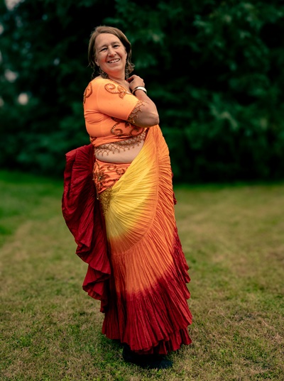 Anita Griebl steht seitlich im bunten Bauchtanzrock, hebt den Stoff zur Schulter und lächelt – ein Ausdruck ihrer weiblichen, freien Energie.