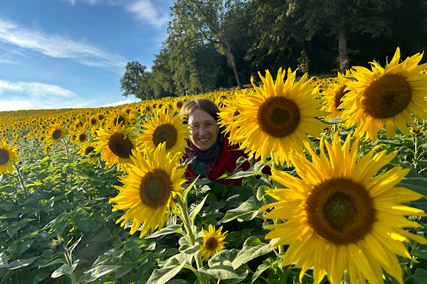 Anita steht unter hohen Sonnenblumen, halb verborgen und sanft hervorschauend – ein zarter Moment, der zeigt, wie Begleitung dir Zeit, Geld und Nerven sparen kann.