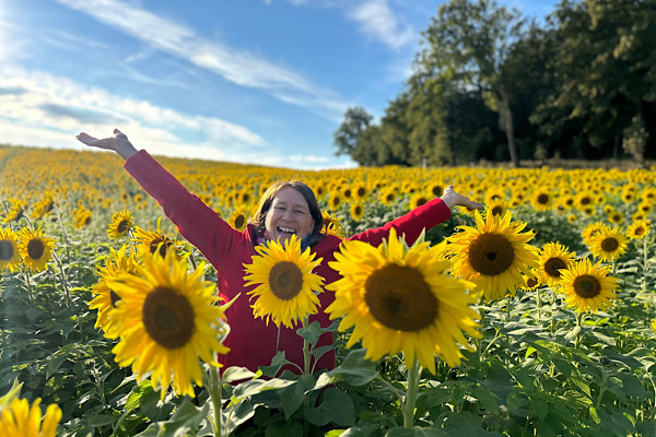 Anita steht in einem Sonnenblumenfeld und streckt die Arme weit aus – ein Moment von Freiheit, der zeigt, wie Begleitung dir Zeit, Geld und Nerven sparen kann.