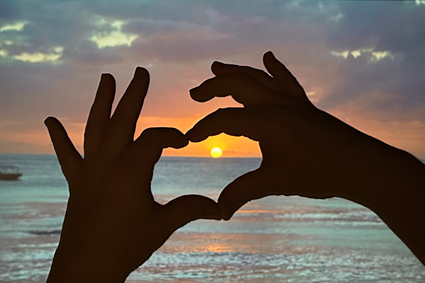 Zwei Hände formen ein Herz vor einem Meer mit Sonnenaufgang.