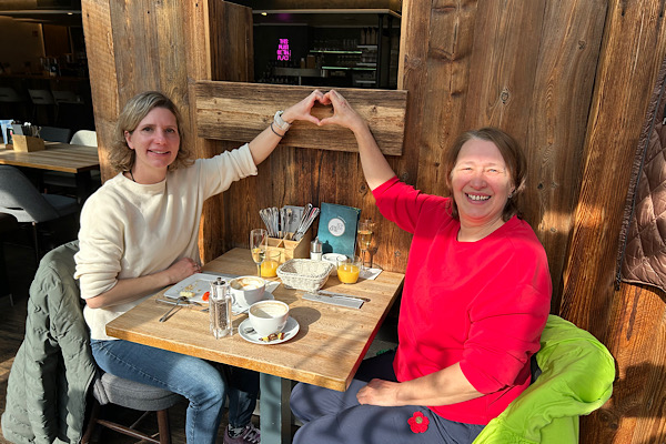 Anita und Martina sitzen beim Frühstück und formen gemeinsam mit ihren Händen ein Herz in der Luft.