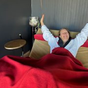 Anita in weißem Bademantel liegt entspannt auf einer Schaukelliege, mit roter Decke zugedeckt, lächelt und streckt die Arme nach oben.