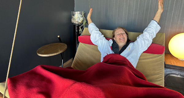 Anita in weißem Bademantel liegt entspannt auf einer Schaukelliege, mit roter Decke zugedeckt, lächelt und streckt die Arme nach oben.