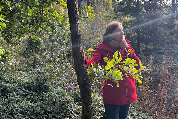 Anita Griebl steht im Wald, die Hand am Kinn, schaut nachdenklich in die Ferne, während ein Lichtstrahl ihr Gesicht berührt.