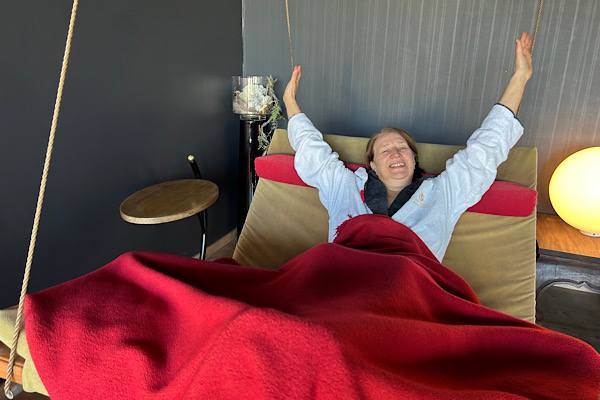 Anita liegt in einer Schaukelliege im weißen Bademantel mit roter Decke und streckt fröhlich die Arme nach oben.