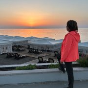 Anita Griebl steht seitlich am Strand im warmen Licht des Sonnenaufgangs und blickt ruhig in die Ferne – Symbol für Orientierung, Klarheit und inneren Weg.