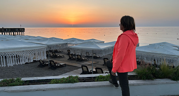 Anita Griebl steht seitlich am Strand im warmen Licht des Sonnenaufgangs und blickt ruhig in die Ferne – Symbol für Orientierung, Klarheit und inneren Weg.