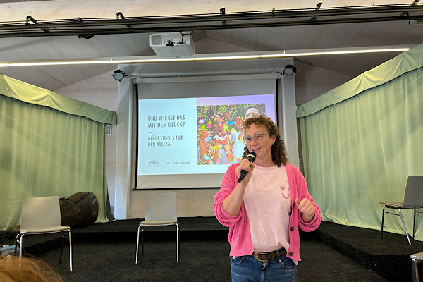 Carola Hanusch während ihres Glücksvortrags, stehend vor ihrer PowerPoint‑Präsentation.